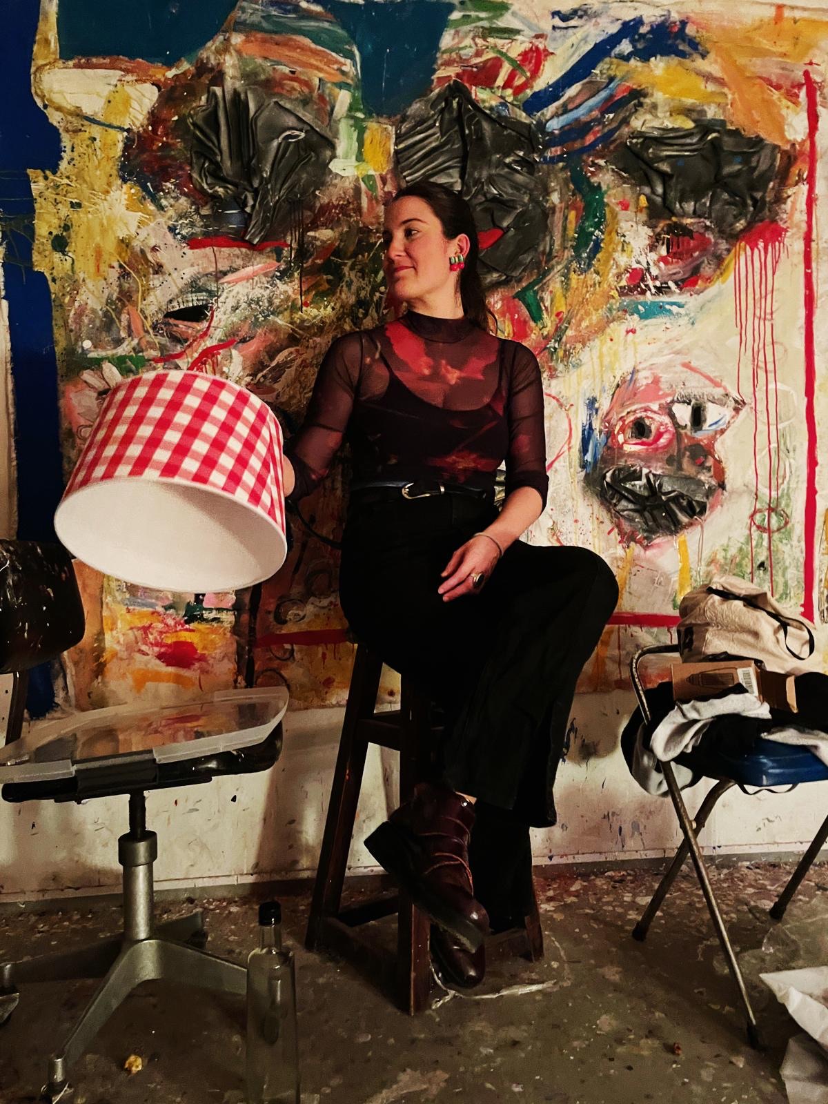Popi Domínguez sentada en un banco junto a una pantalla de lámpara, frente a una pintura